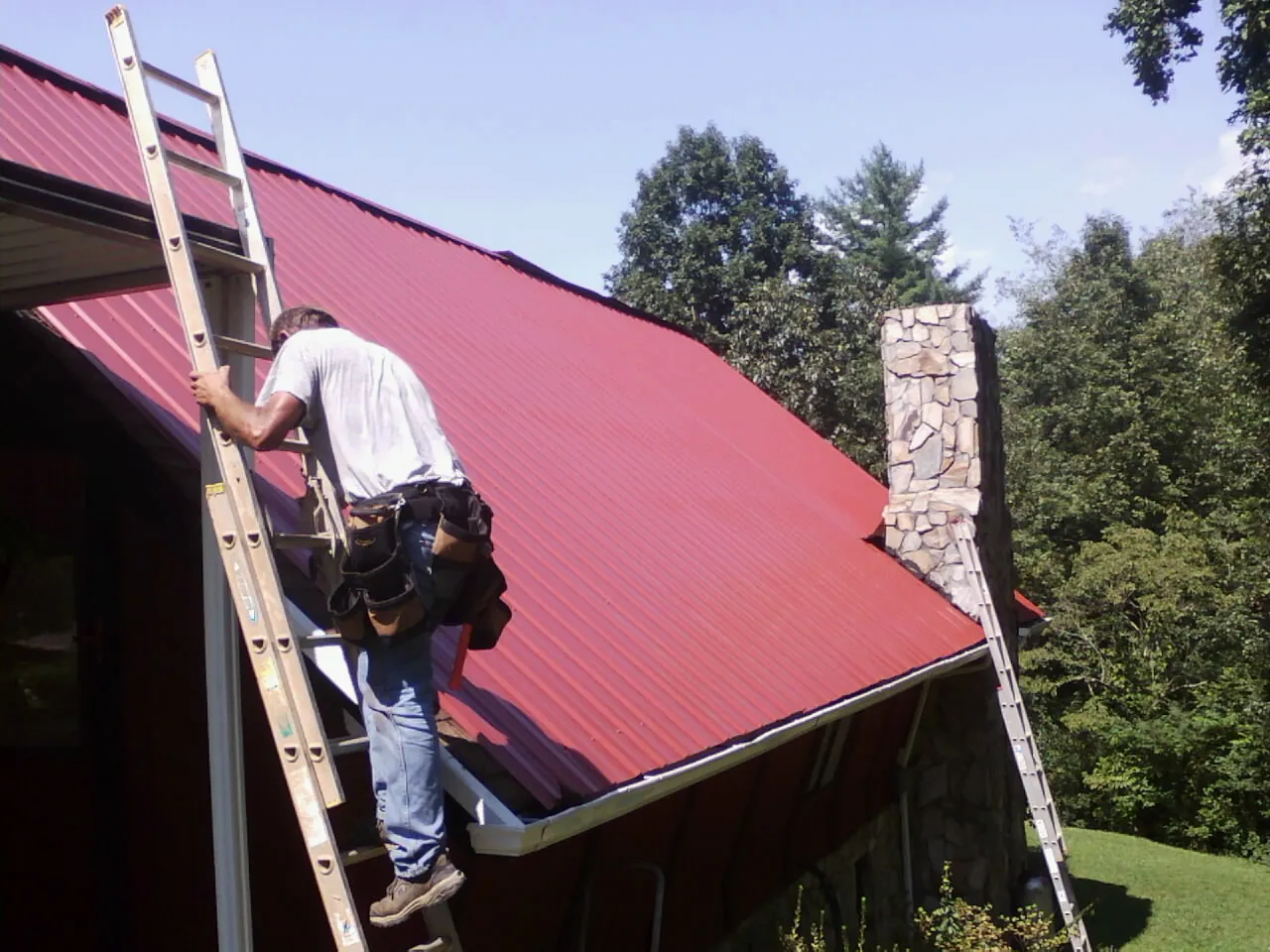 Roofer on a ladder beside a red metal roof and stone chimney — Soffit & Fascia Repair by Roofing Brothers in Gap